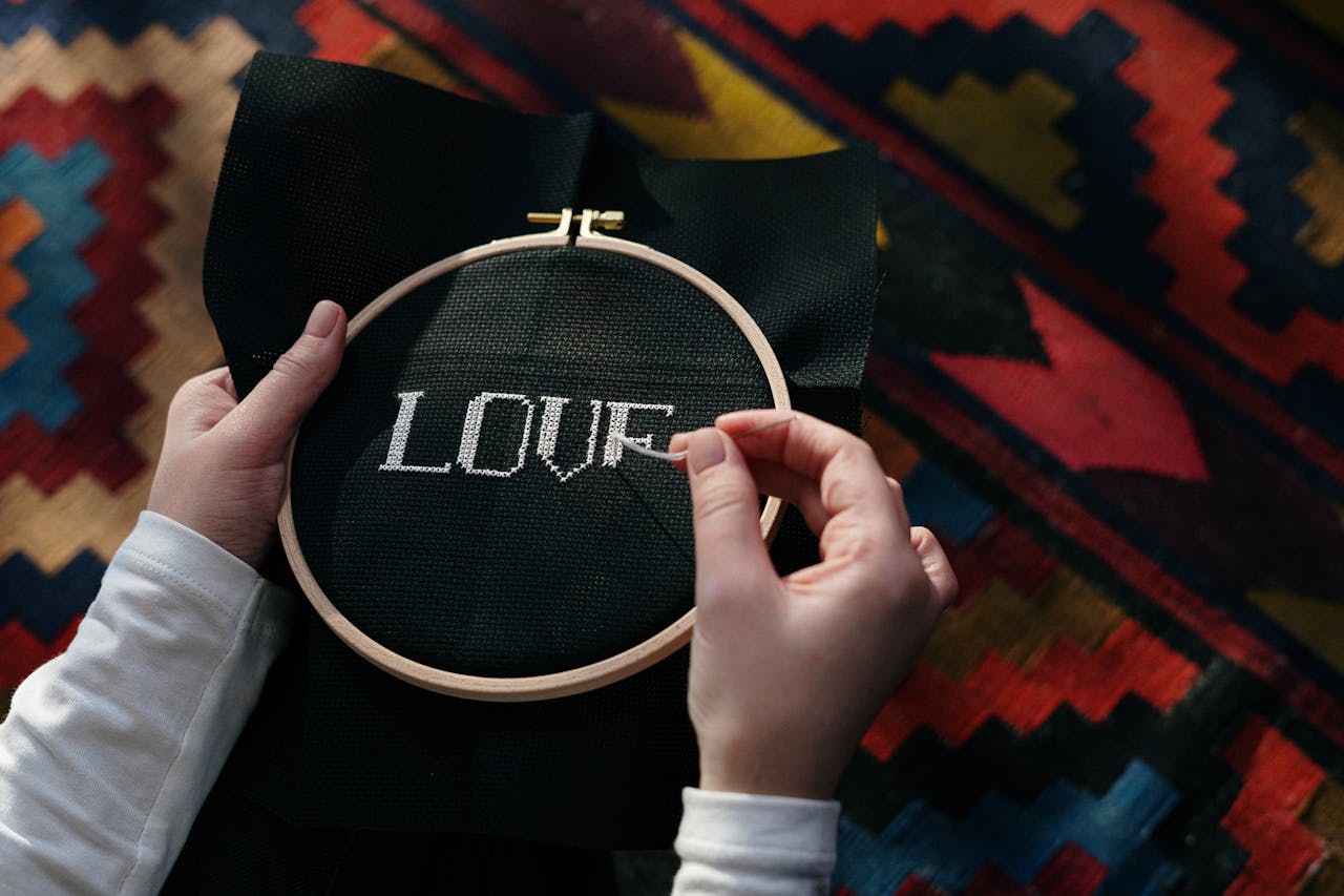 Hands embroidering 'LOVE' on black fabric using a wooden hoop, close-up view.